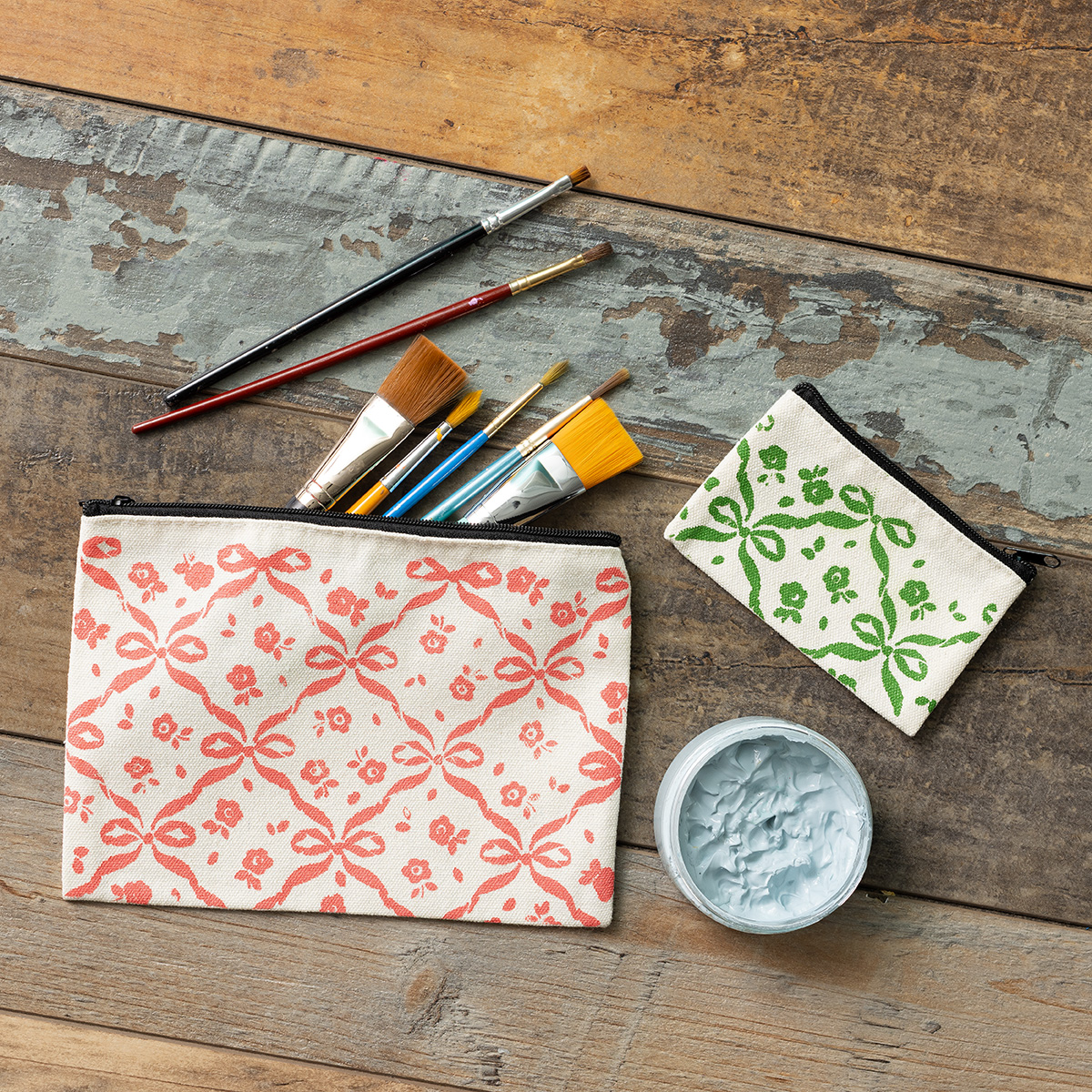 A flat lay of paintbrushes, a jar of blue paint, and two zippered pouches with hand-stamped patterns—one larger with a red design, one smaller with a green design—on a rustic wooden surface.