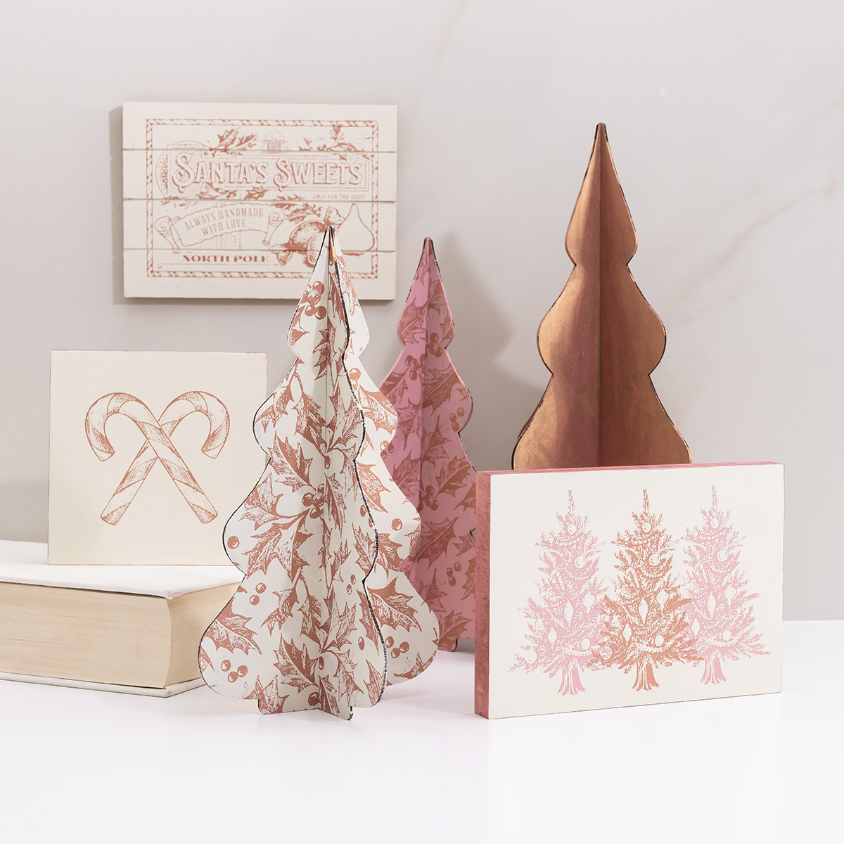 A display of holiday decorations includes illustrated block signs and 3D paper trees in pink, cream, and brown tones, featuring Christmas-themed designs like candy canes, trees, and script text.