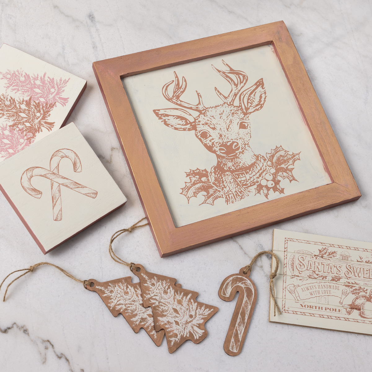 A framed sketch of a reindeer with holly, two small cards with holiday designs, three brown tree-shaped ornaments, a candy cane ornament, and a vintage-style "Santa's Sweet Shoppe" tag on a marble surface.
