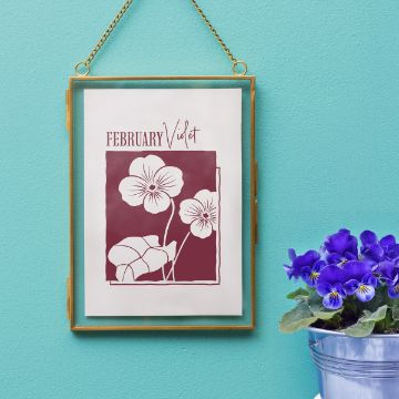 Show details for Violet (February) A framed illustration labeled "February Violet" hangs on a teal wall, featuring violet flowers in maroon and white. Next to it sits a potted violet plant with vibrant purple blooms.