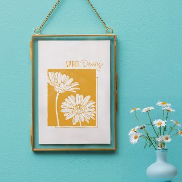 Show details for Daisy (April) A gold-framed picture hangs on a turquoise wall, displaying an illustration of two daisies with the text "April Daisy" above them. Next to it, a small blue vase holds fresh white daisies.