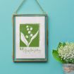 A framed artwork of white lily of the valley flowers with green leaves and the words "May Lily of the Valley" hangs on a turquoise wall. A vase with fresh lily of the valley sits nearby.