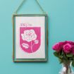 A framed print with a pink illustration of a rose and the text "June Rose" hangs on a turquoise wall. A vase with pink roses sits nearby on a table.