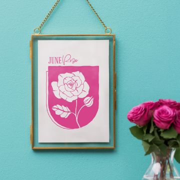 Show details for Rose (June) A framed print with a pink illustration of a rose and the text "June Rose" hangs on a turquoise wall. A vase with pink roses sits nearby on a table.