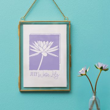 Show details for Water Lily (July) A gold-framed picture of a white water lily with "July Water Lily" written below hangs on a turquoise wall; nearby is a glass vase with two blooming water lilies.