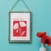 A gold-framed picture hangs on a blue wall, displaying red and white poppy flower art labeled "AUGUST Poppy." A small vase with red poppies sits below on the right.