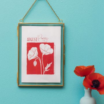 Show details for Poppy (August) A gold-framed picture hangs on a blue wall, displaying red and white poppy flower art labeled "AUGUST Poppy." A small vase with red poppies sits below on the right.