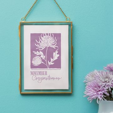 Show details for Chrysanthemum (November) A framed artwork featuring a purple illustration of chrysanthemums labeled "November Chrysanthemum" hangs on a teal wall, with a vase of light purple chrysanthemums beside it.