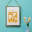 A gold-framed artwork hangs on a blue wall, featuring daffodil illustrations and the text "December Narcissus" in orange. Nearby, a light blue vase holds three white narcissus flowers.