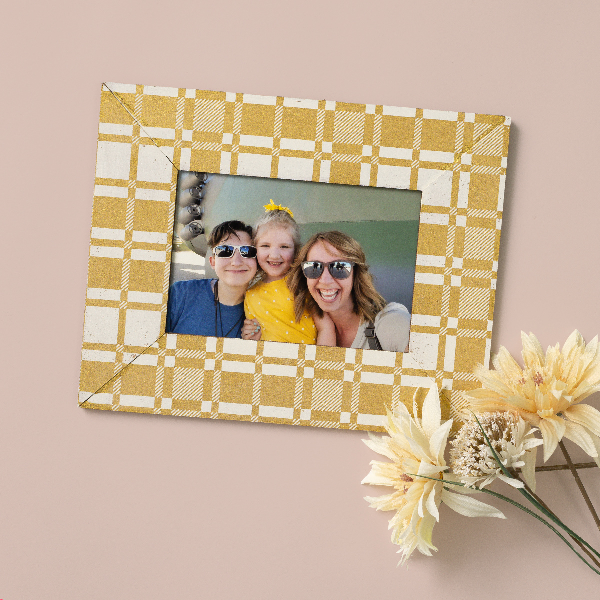 A framed photo of a smiling woman and two children, all wearing sunglasses. The frame has a yellow plaid pattern and sits on a light pink surface beside a bunch of pale yellow flowers.