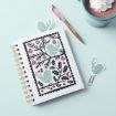 A spiral notebook with a cover featuring green birds and holly branches, a pink and gold pen, a mint mug of hot chocolate with whipped cream, and two bird-shaped paper clips on a pale blue surface.