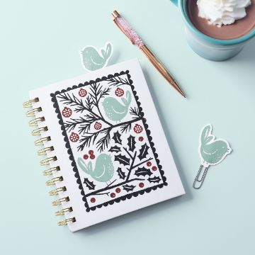 Show details for Birds & Branches A spiral notebook with a cover featuring green birds and holly branches, a pink and gold pen, a mint mug of hot chocolate with whipped cream, and two bird-shaped paper clips on a pale blue surface.