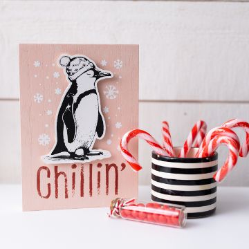 Show details for Chillin' A card with a drawing of a penguin wearing a winter hat and the word "chillin'" stands next to a striped cup filled with candy canes and a small glass jar of red candy sticks on a white surface.