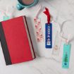 A red book, four colorful bookmarks with festive designs, and a teal mug of hot chocolate are arranged on a white marble surface.