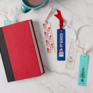 Show details for I'm Booked A red book, four colorful bookmarks with festive designs, and a teal mug of hot chocolate are arranged on a white marble surface.