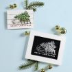 Two framed “Merry Christmas” cards featuring a sled carrying a tree, surrounded by pine sprigs and gold jingle bells on a light blue surface.