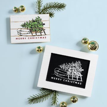 Show details for Christmas Sled Two framed “Merry Christmas” cards featuring a sled carrying a tree, surrounded by pine sprigs and gold jingle bells on a light blue surface.