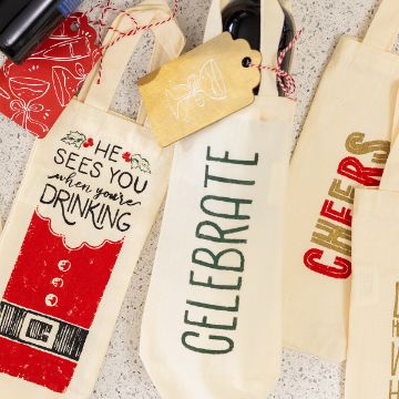 Show details for Cheers Three festive wine bottle bags on a speckled surface; one reads "He sees you when you're drinking" with a Santa design, another says "CELEBRATE," and the third says "CHEERS" in red and gold letters.