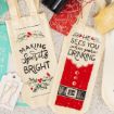 Two festive wine bags on a counter: one has holly and reads "Making Spirits Bright," the other looks like a Santa suit and says "He sees you when you're drinking." Gift tags and part of a bottle are also visible.