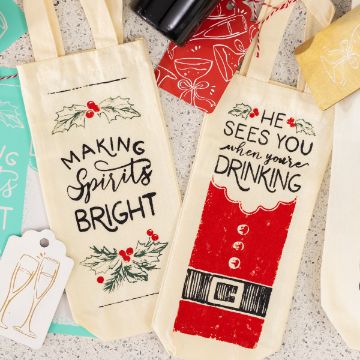 Show details for When You're Drinking Two festive wine bags on a counter: one has holly and reads "Making Spirits Bright," the other looks like a Santa suit and says "He sees you when you're drinking." Gift tags and part of a bottle are also visible.