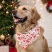 A golden retriever wearing a festive white bandana with red holiday designs sits indoors near a decorated Christmas tree and twinkling lights, looking happy with its tongue out.