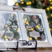 A frosty, wooden-framed glass panel decorated with white drawings of Christmas ornaments, set against a background of blurred yellow holiday lights and a decorated Christmas tree.