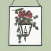 Framed illustration of a vintage lantern with a lit candle inside, decorated with pine branches, pinecones, and a red bow. The frame hangs from a chain against a light green background.