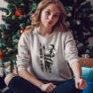 A young woman sits in front of a decorated Christmas tree, wearing a light-colored sweatshirt with a playful Santa Claus and the text "I SAW THAT." She looks at the camera with a relaxed expression.