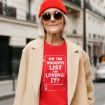 Smiling older woman in sunglasses and a red beanie wears a red shirt with white text that reads, “On the naughty list and loving it!” She stands outdoors in a city street, wearing a beige coat.