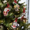 Four fabric Christmas ornaments—a Santa, an elf, a reindeer, and a penguin holding a present—hang on a decorated Christmas tree with twinkling white lights.