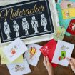 A person arranges festive greeting cards featuring illustrated mice, a snowman, and holiday themes next to a framed "The Nutcracker" sign with nutcracker figures displayed in white on a black background.