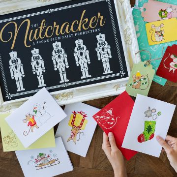 Show details for Merry Mouse A person arranges festive greeting cards featuring illustrated mice, a snowman, and holiday themes next to a framed "The Nutcracker" sign with nutcracker figures displayed in white on a black background.