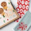 A tray with two drinks sits on a festive cloth with holiday icons. Nearby are two holiday-themed napkins: one red with white ovals and Christmas symbols, and one green and white striped with a large bow design.