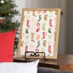 A framed print with various colorful Christmas stockings is displayed on a wooden table. The background features a decorated Christmas tree and red and white pillows in the foreground.
