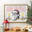 Framed vintage-style Merry Christmas print with Santa Claus, candles, and festive text on a white wall. Nearby are wrapped gifts, a small decorated Christmas tree, and red and white lanterns.
