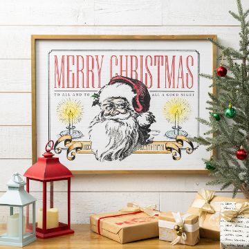 Show details for Santa's Wish Framed vintage-style Merry Christmas print with Santa Claus, candles, and festive text on a white wall. Nearby are wrapped gifts, a small decorated Christmas tree, and red and white lanterns.
