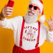 A man dressed as Santa Claus in red suspenders and sunglasses takes a selfie, sticking out his tongue and making a rock hand gesture, against a bright yellow background. His shirt reads "LET'S GET FROSTED" with a snowman graphic.