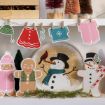 A festive holiday scene featuring gingerbread people, a snowman plate with a red bird, mini Christmas trees, and a garland with tiny paper clothes like hats, mittens, shirts, and dresses hanging above.