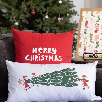 Show details for Elf Buddies Two festive pillows on a couch; one red with the words "Merry Christmas," and one white with an illustration of Santa decorating a tree. A Christmas tree and framed holiday-themed print are in the background.