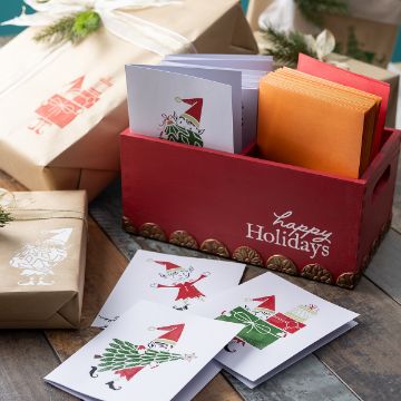 Show details for Santa's Elves A red box labeled "Happy Holidays" holds festive cards and envelopes. Three cards with holiday-themed gnome illustrations are displayed on a wooden surface beside wrapped gifts with gnome designs.