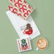 A festive flat lay with a red and beige oven mitt, a white tea towel featuring gingerbread cookies and "Let's Get Baked" text, a holiday-themed coaster, and a cup of latte with heart-shaped latte art.
