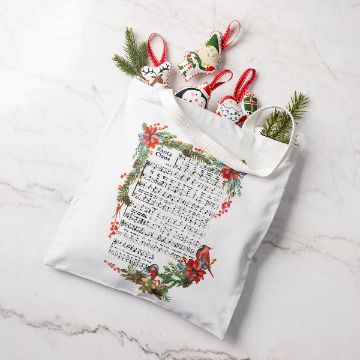 Show details for Heat Transfer Tote A white tote bag decorated with sheet music and holiday greenery holds Christmas ornaments shaped like Santa, a gingerbread man, and more, with sprigs of evergreen, all set on a marble surface.