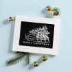 Framed card with a white sketch of a sled carrying a Christmas tree and the words “Merry Christmas.” Surrounded by gold jingle bells and a pine branch on a light blue background.