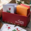A red wooden box labeled "happy Holidays" holds festive cards with elves and orange envelopes, set on a wooden table with some cards spread out nearby and wrapped gifts in the background.