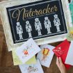 A framed Nutcracker sign sits on a wooden surface, surrounded by holiday cards featuring festive illustrations like a snowman, a mouse with cheese, a gift, and a sled. A hand holds a red card with a white mouse design.