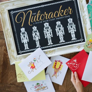 Show details for Regency Artisan Add-Ons A framed Nutcracker sign sits on a wooden surface, surrounded by holiday cards featuring festive illustrations like a snowman, a mouse with cheese, a gift, and a sled. A hand holds a red card with a white mouse design.