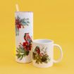 A tall tumbler with a straw and a mug, both white with festive designs featuring red cardinals, red flowers, and green foliage, are set against a solid yellow background.