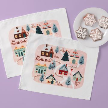 Show details for The North Pole Heat Transfer Two placemats with a whimsical North Pole map design showing houses, trees, and labeled landmarks, next to a white plate holding four star-shaped decorated cookies on a light purple surface.