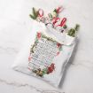 A white tote bag decorated with Christmas sheet music and festive illustrations holds evergreen sprigs and various Christmas ornaments on a white marble surface.