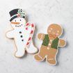 Two holiday cookies shaped like a snowman and a gingerbread person rest on a white marble surface. The snowman wears a black hat, holds a candy cane, and the gingerbread person has green plaid details and a red bow tie.